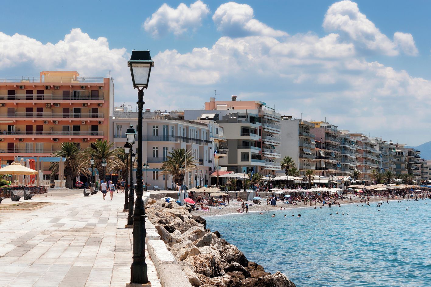 A coastal promenade with a stone walkway, a beach with umbrellas, and multi-story buildings in the background.