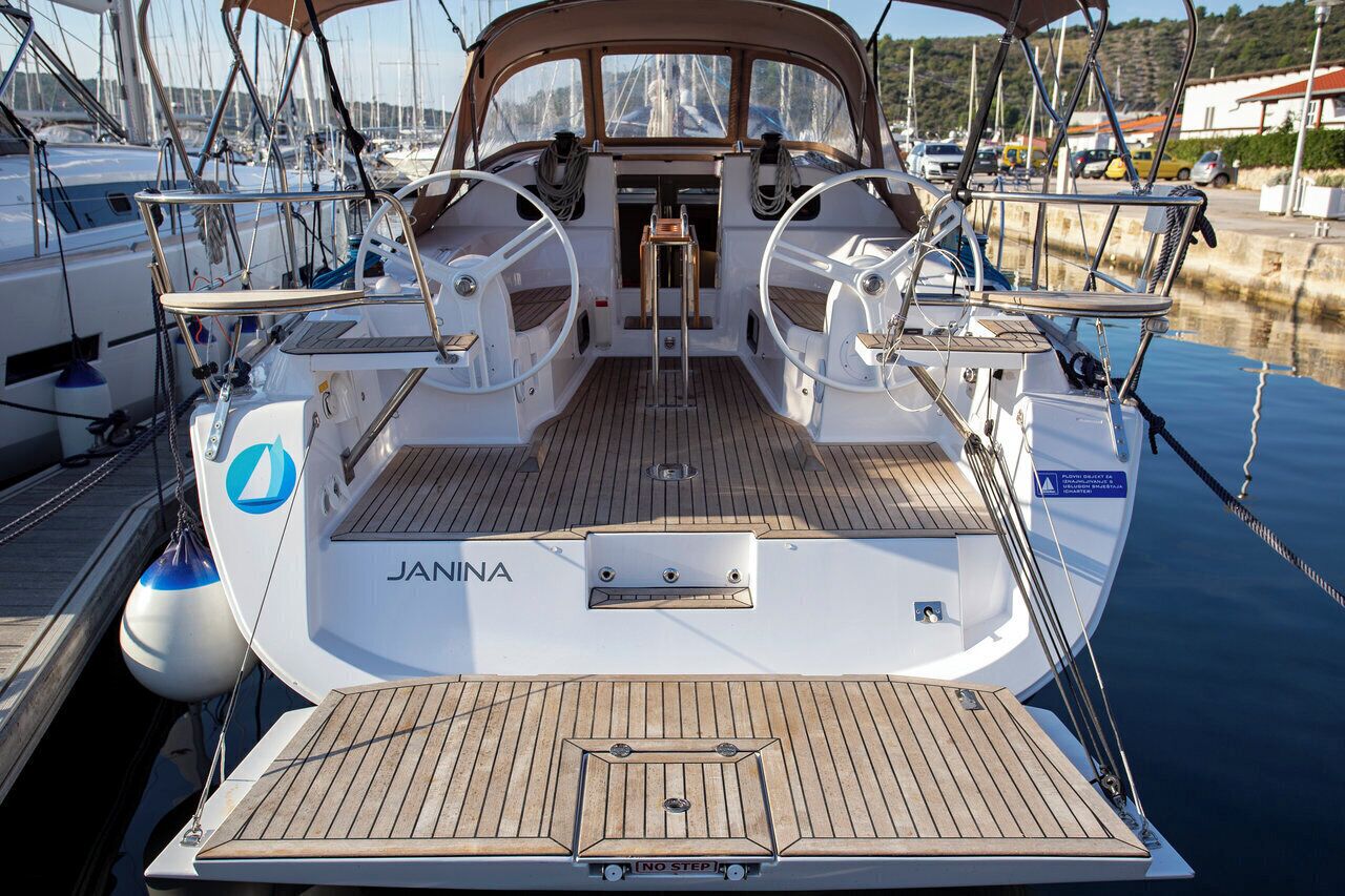 A sailboat named JANINA docked at a marina with a wooden deck, steering wheel, and a small cabin area.