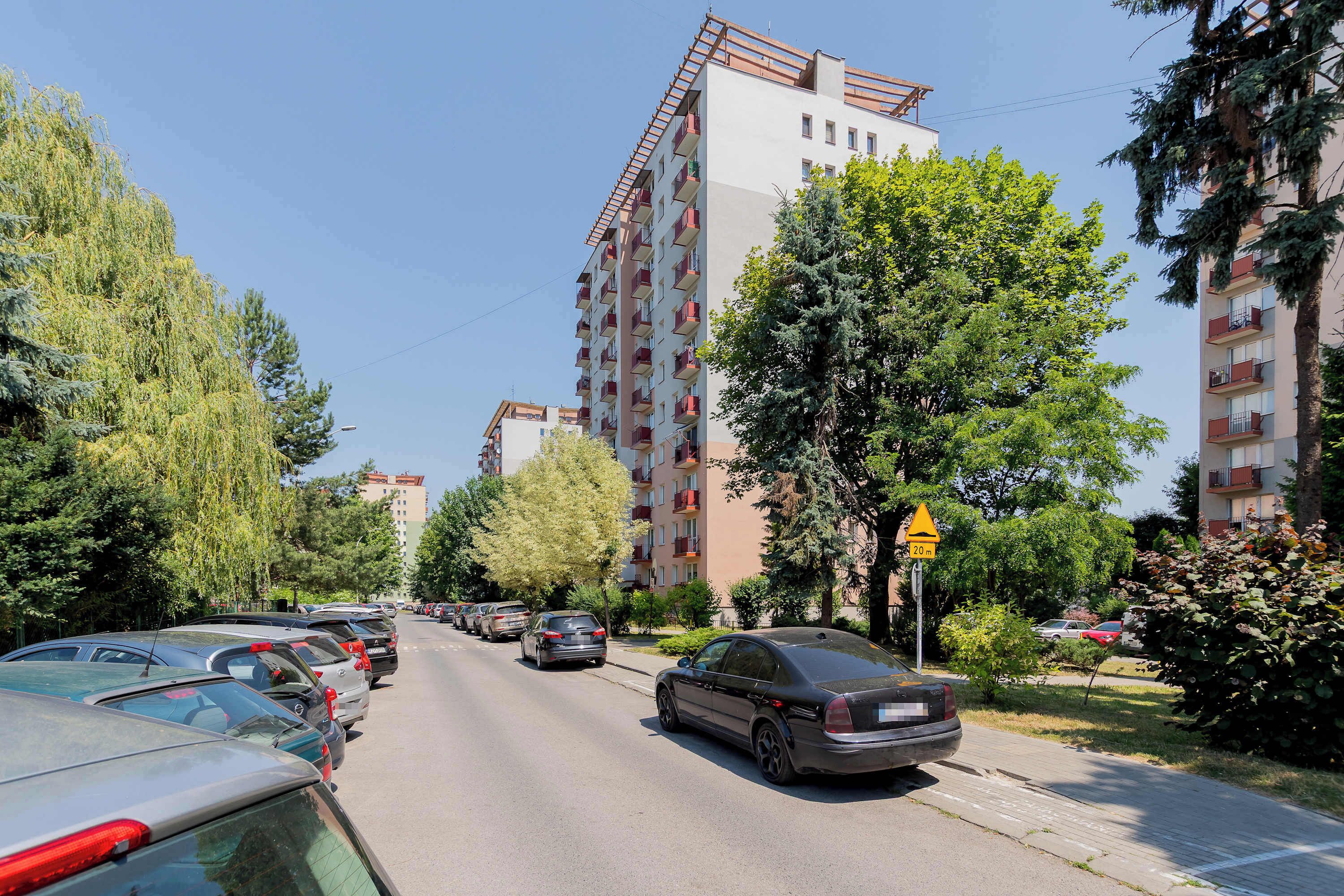 A row of parked cars along a street lined with multi-story residential buildings, trees, and a clear sky.