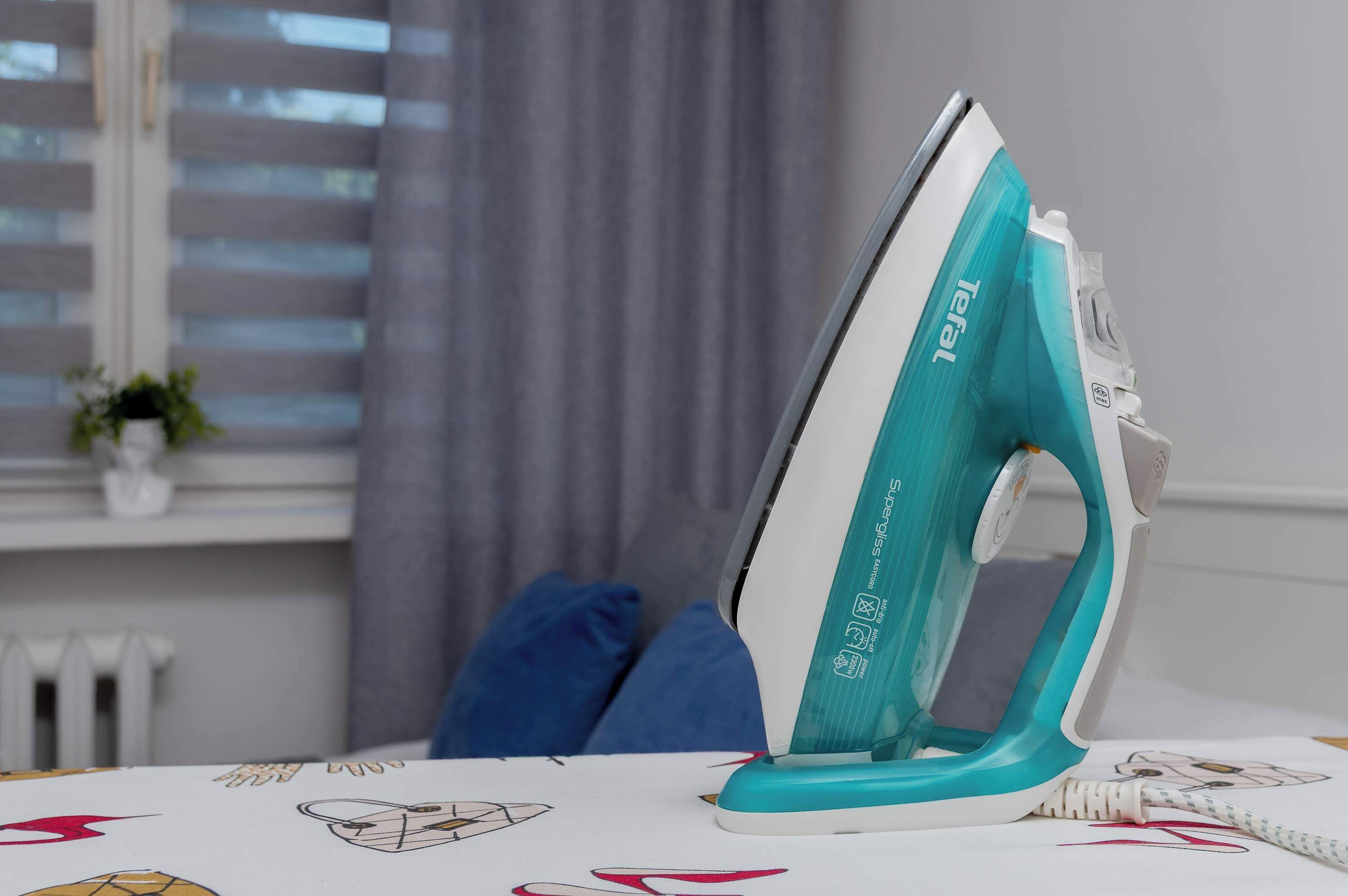 An ironing board with an iron, a window with blinds, and a plant in the background.