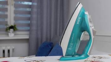An ironing board with an iron, a window with blinds, and a plant in the background.