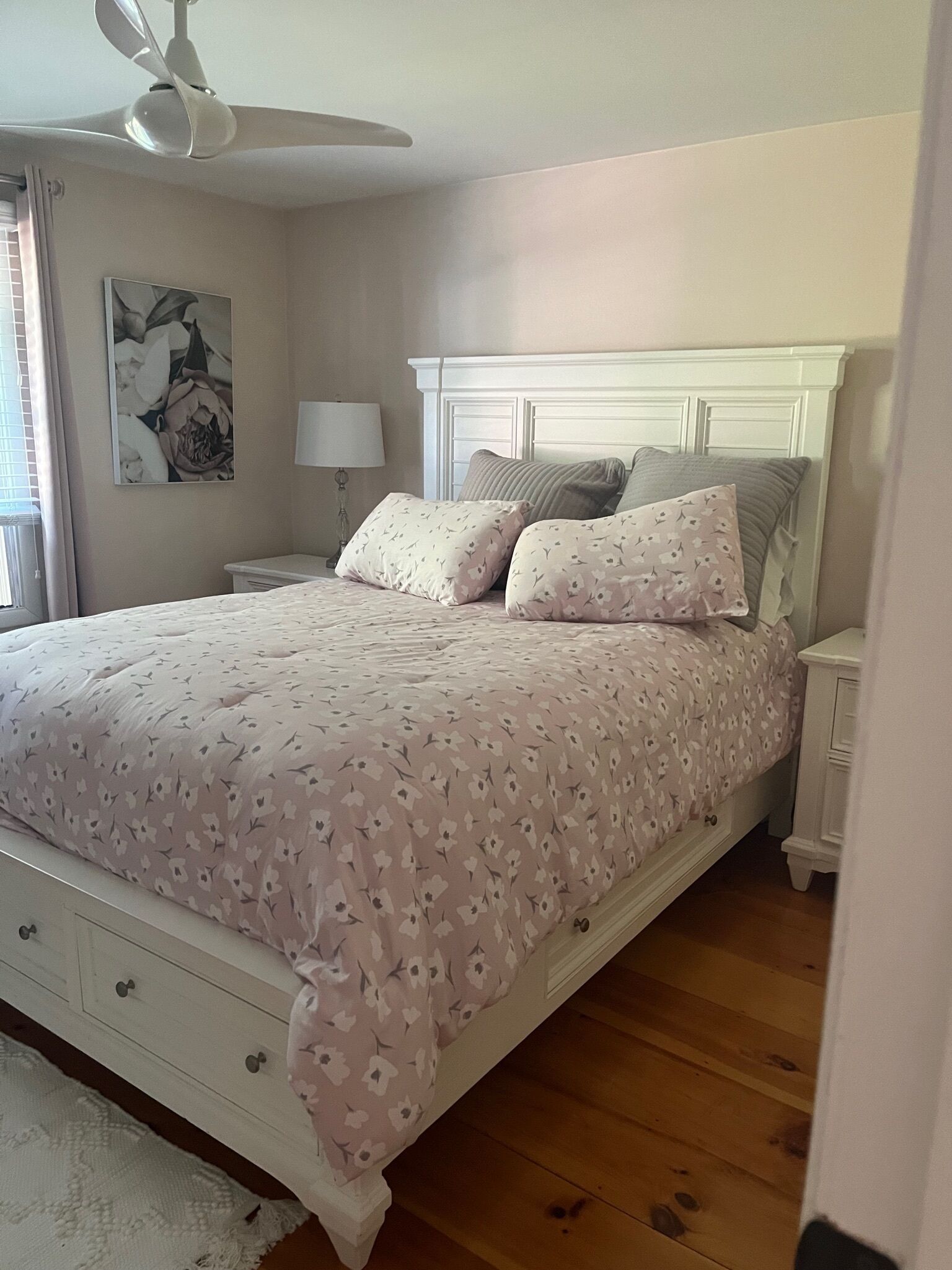 A bedroom with a bed, a nightstand, a lamp, a ceiling fan, and a wall with a floral painting.