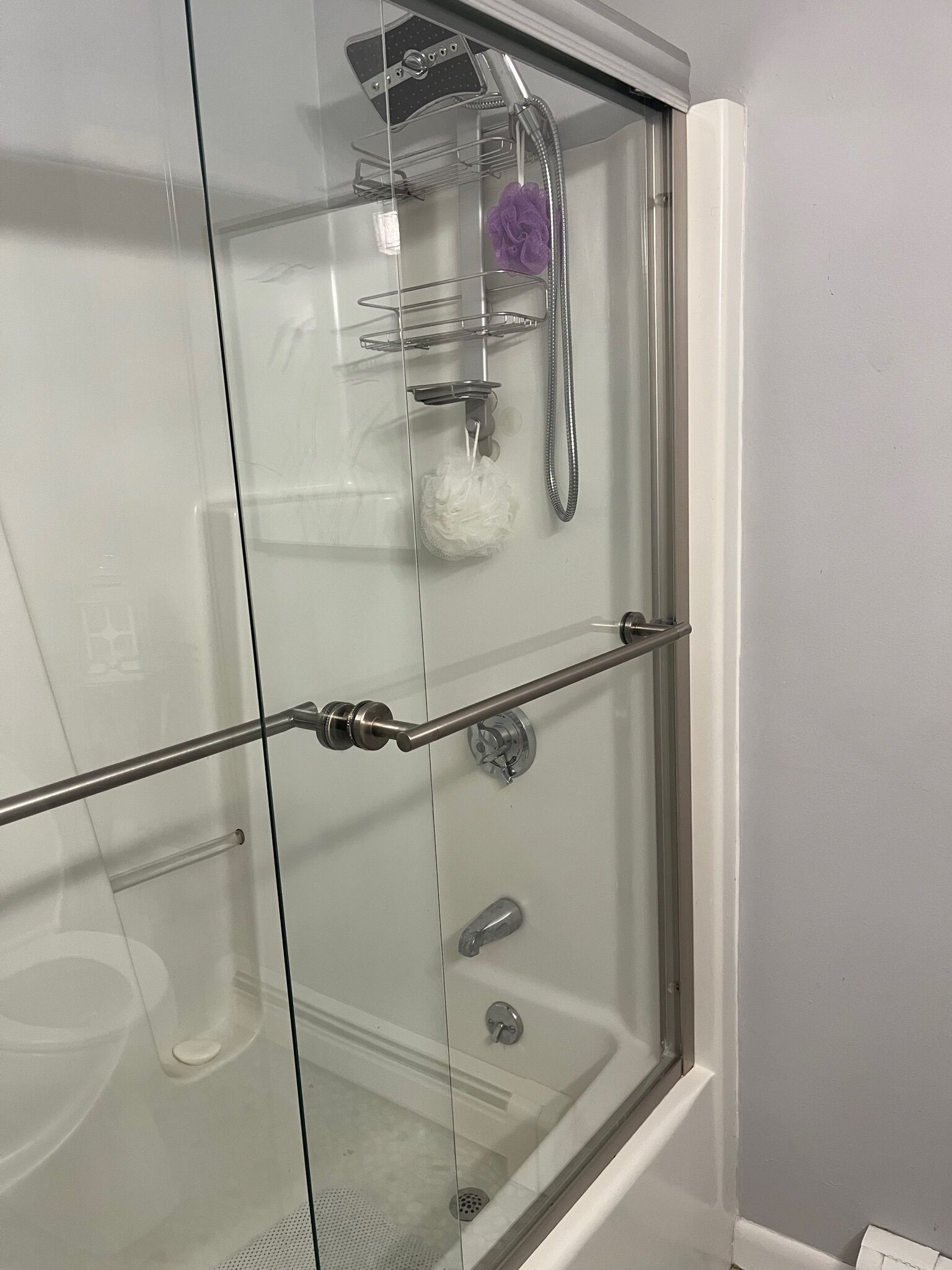 A glass-enclosed shower with a handheld showerhead, a handheld shower attachment, and a white bath mat.