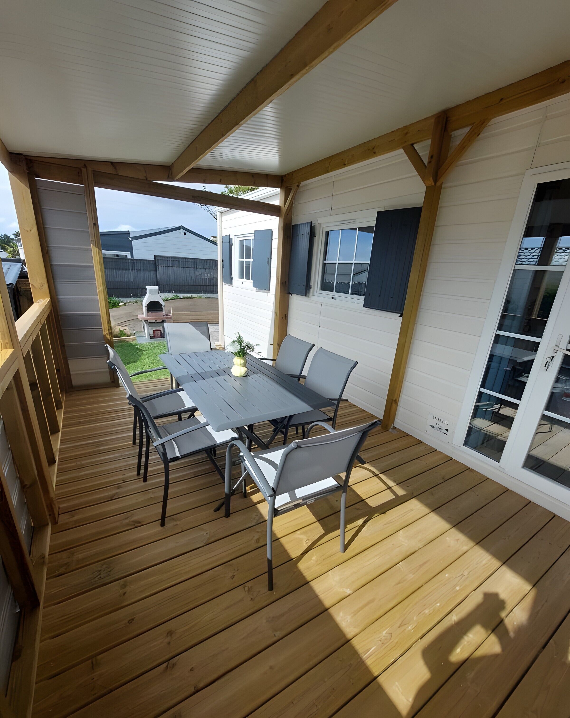 Een overdekt terras met een houten terras, een tafel met stoelen en uitzicht op een huis met een raam en een deur.