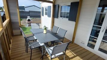 Een overdekt terras met een houten terras, een tafel met stoelen en uitzicht op een huis met een raam en een deur.