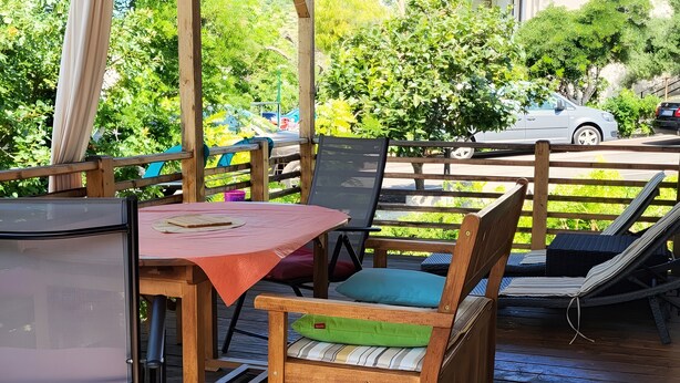 A wooden deck with a table and chairs, a grill, and a view of a street with cars and greenery.