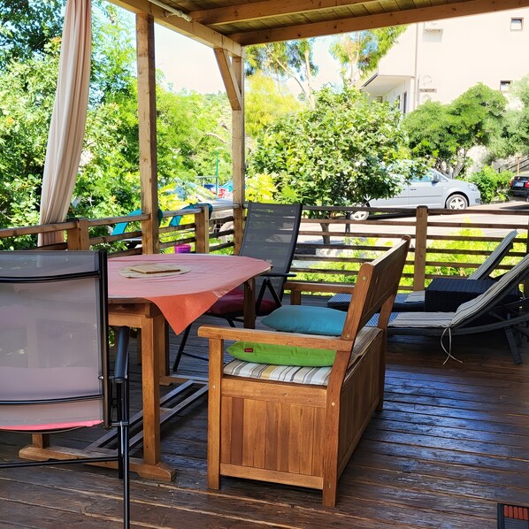 Eine Holzterrasse mit Tisch, Stühlen, einem Grill und Blick auf eine Straße mit Autos und Grünflächen.