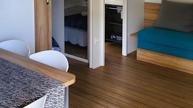 Una habitación con piso de madera, una cama con colchón azul, una silla blanca y una mesa con mantel estampado.