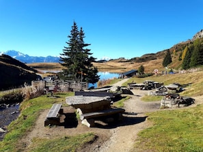 Un área de picnic junto al lago con bancos de piedra, un puente de madera y montañas al fondo.