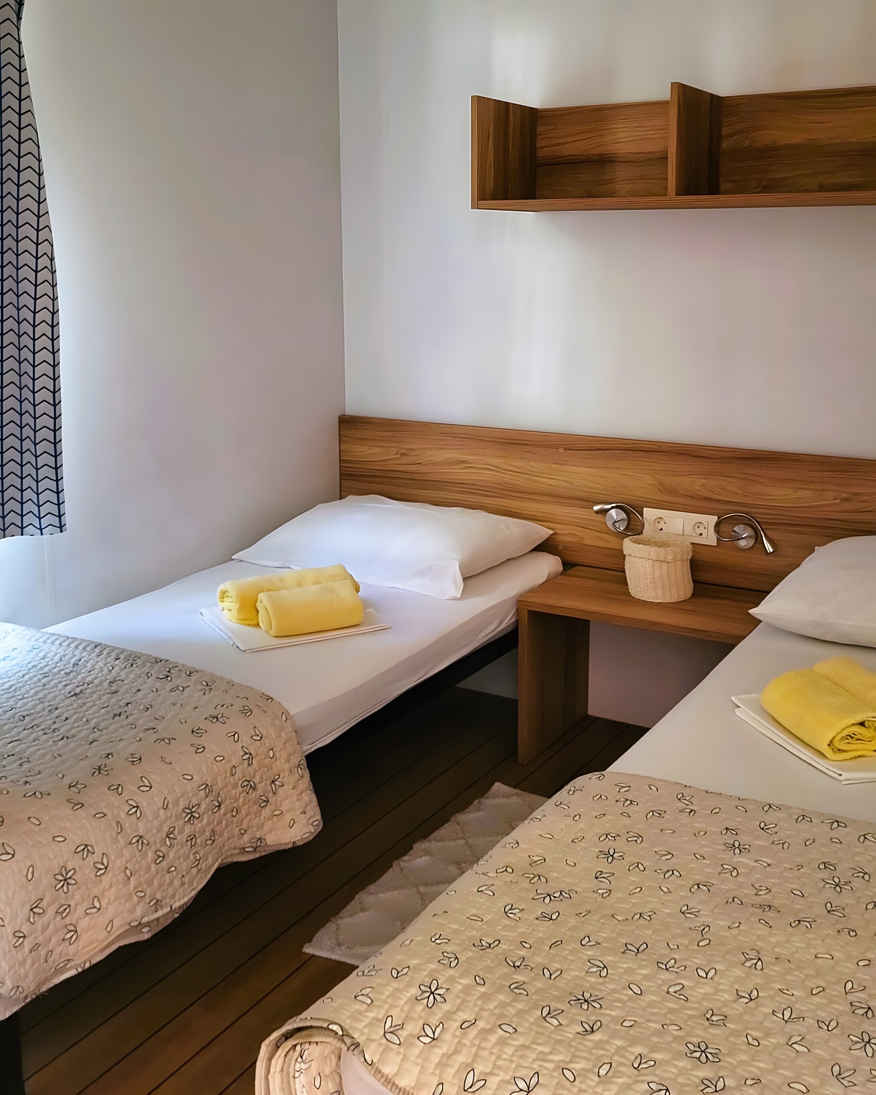 A room with two single beds, a wooden headboard, a bedside table with a lamp, and a wall-mounted shelf.