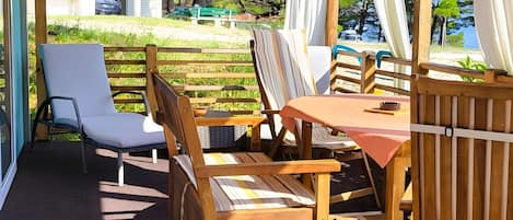 Eine Terrasse im Freien mit Holzmöbeln, darunter Stühle und ein Tisch, und Blick auf Strand und Bäume.