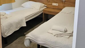 A room with two beds, each with a wooden headboard and white bedding. There is a wooden shelf above the beds and a wall-mounted light fixture.