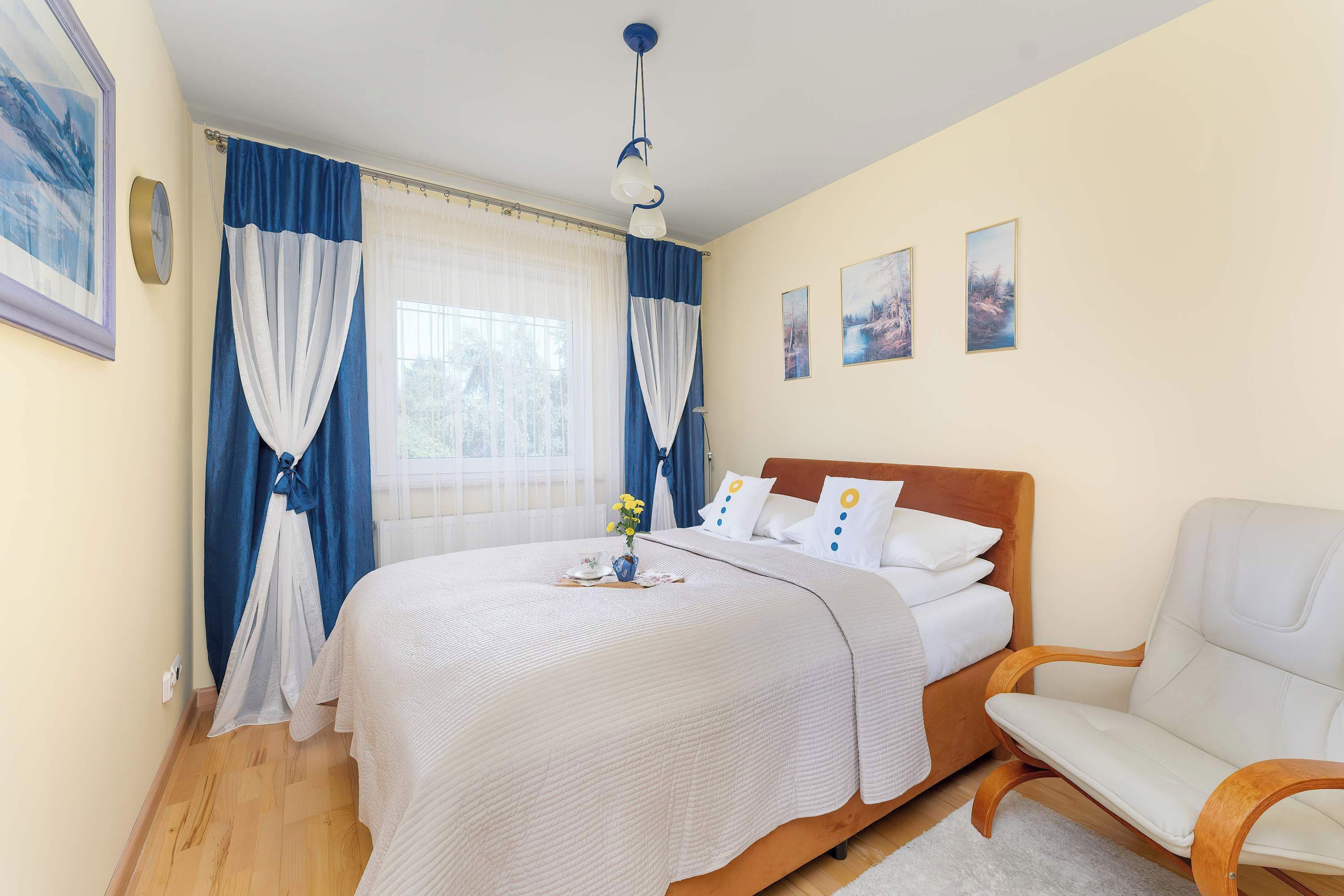 A bright bedroom with a double bed, blue curtains, and light wooden furniture. Large windows bring natural light into the cozy space.