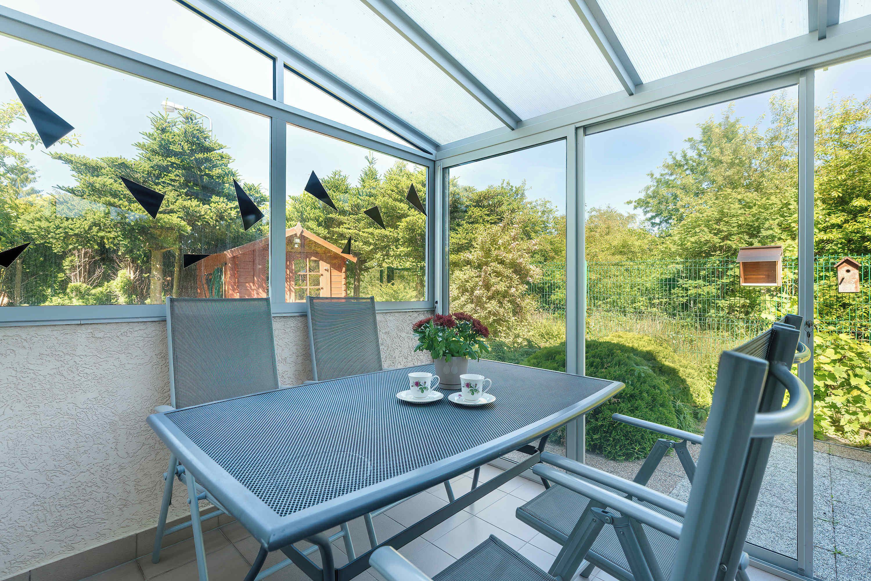 A terrace lounge area furnished with outdoor seating beneath a glass canopy. Green surroundings create a pleasant atmosphere.