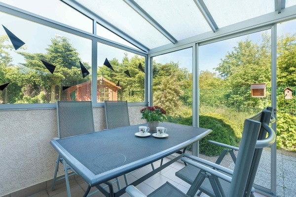 A terrace lounge area furnished with outdoor seating beneath a glass canopy. Green surroundings create a pleasant atmosphere.
