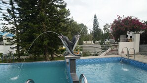 A swimming pool with a dolphin sculpture, surrounded by trees and a building.