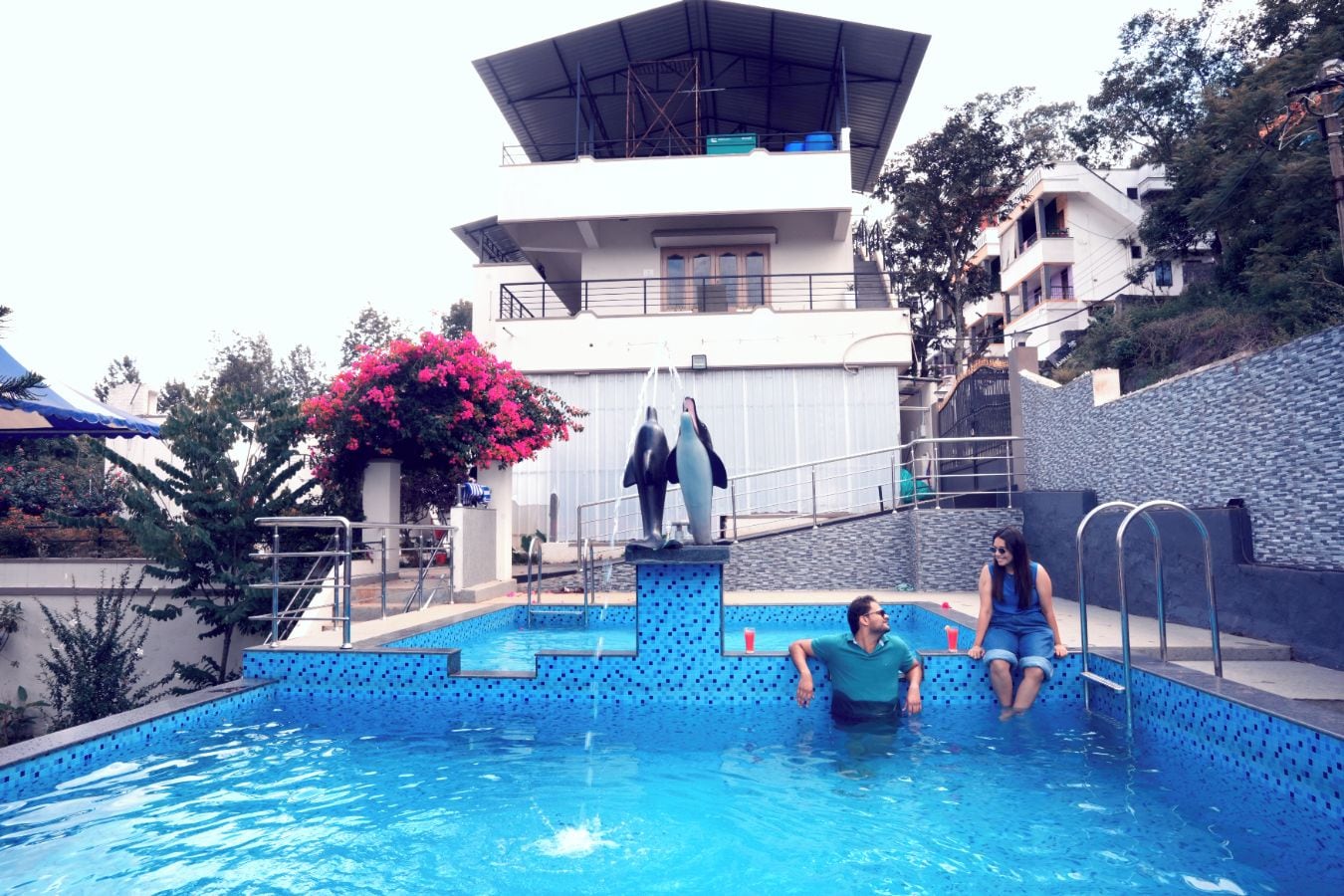 Ein Swimmingpool mit einer Delfinstatue, umgeben von einem modernen Gebäude und einer rosa blühenden Pflanze.