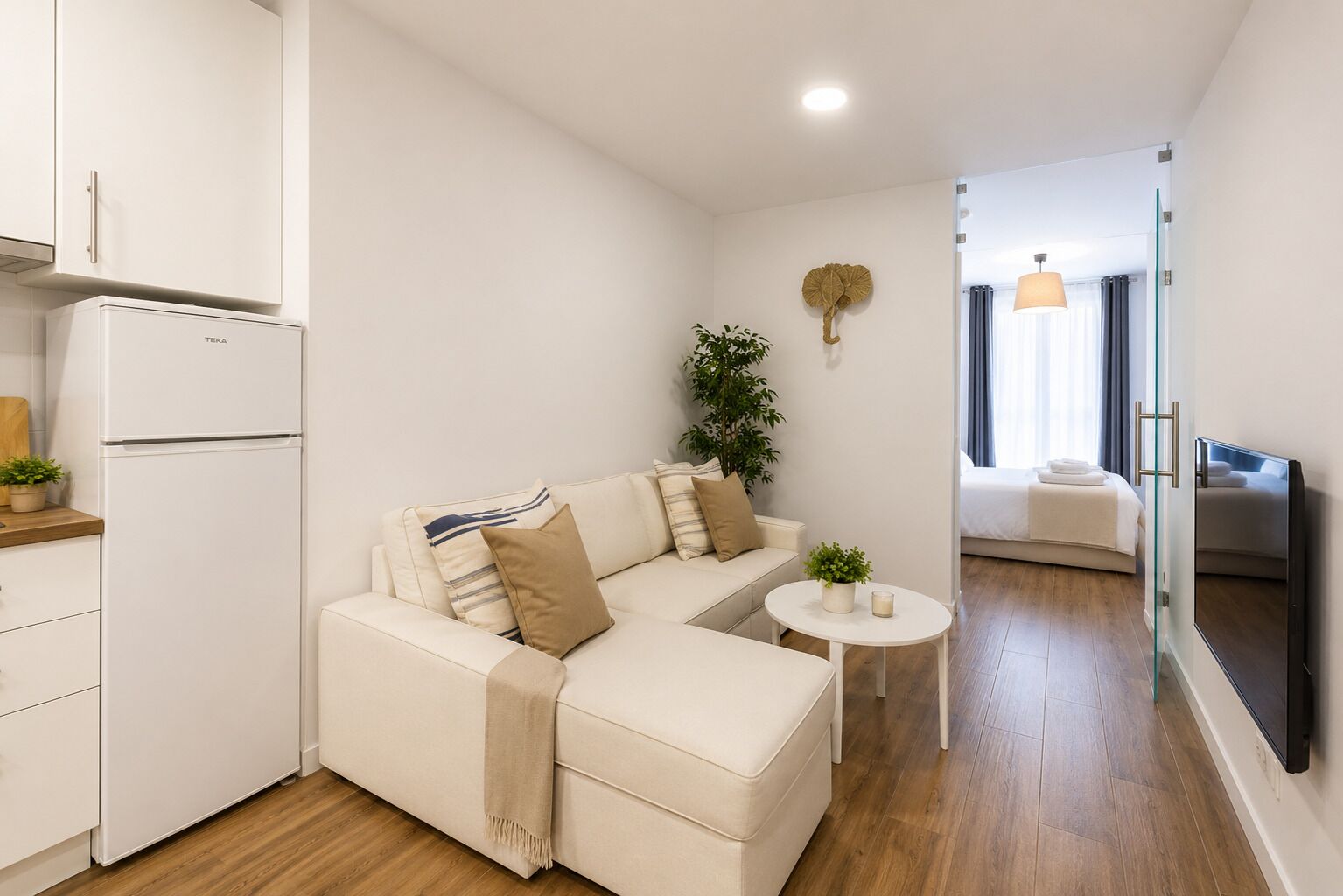 Una habitación moderna y bien iluminada con un sofá, una mesita, una planta y una cama al fondo.