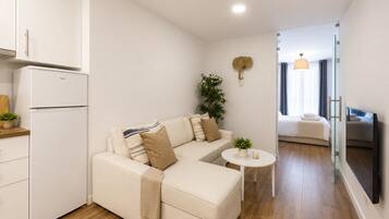 Una habitación moderna y bien iluminada con un sofá, una mesita, una planta y una cama al fondo.