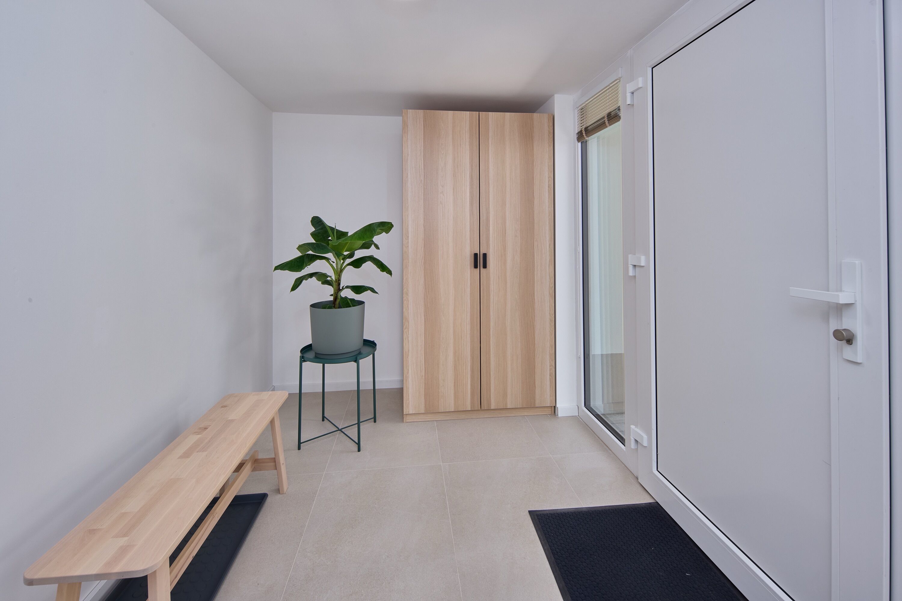 Een gang met een houten bankje, een potplant, een houten garderobe en een glazen deur.