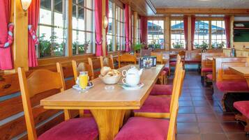 Un ristorante con tavoli e panche in legno, un'ambientazione per la colazione con teiera e succo d'arancia, e ampie finestre con tende.