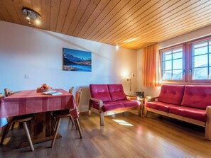 Ein gemütliches Wohnzimmer mit Holzbalkendecke, einem Esstisch mit Stühlen, einem roten Sofa und einem Fenster mit Vorhängen.