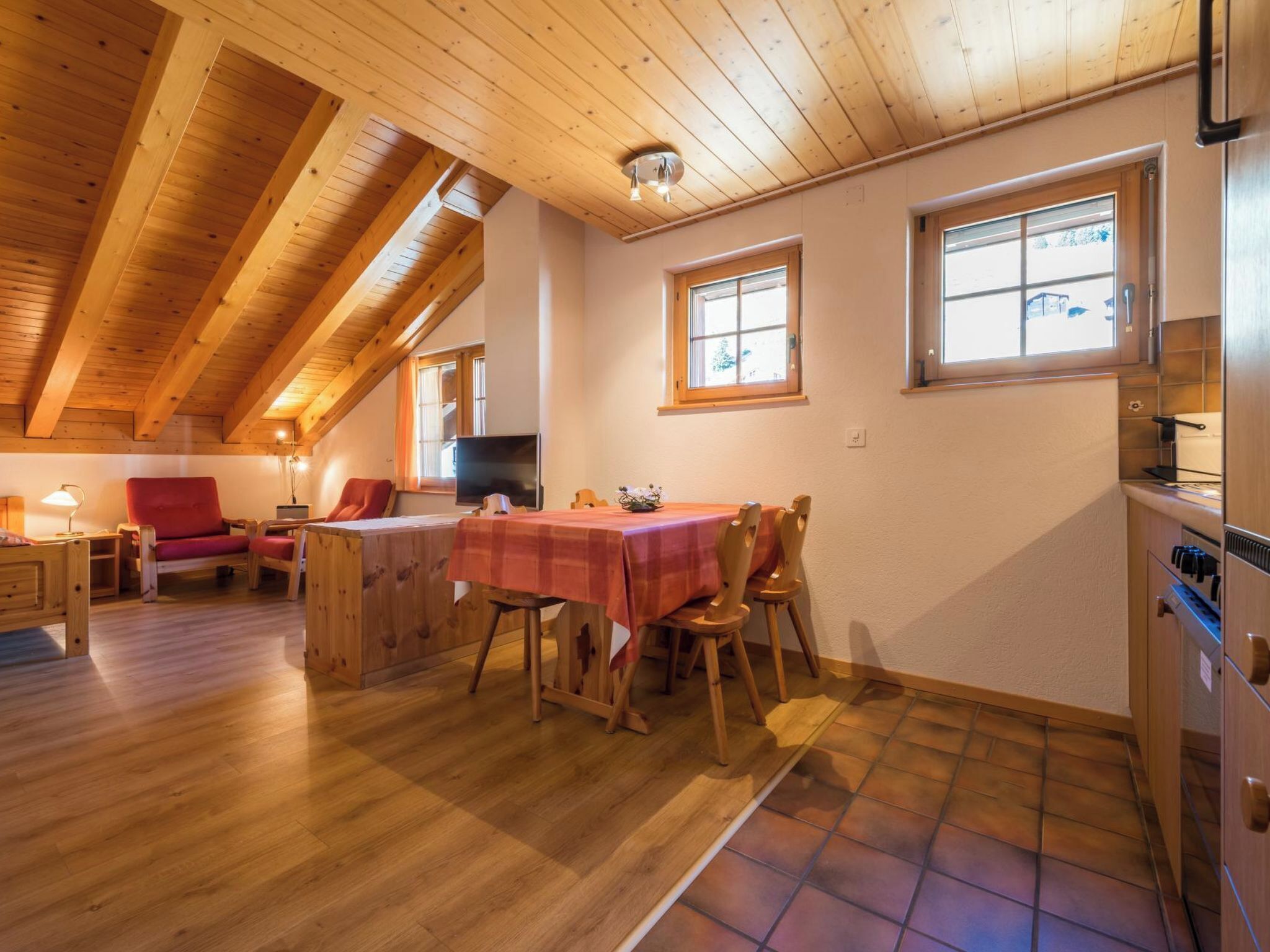A cozy, wooden-floored dining area with a table set for two, a red sofa, and a television.