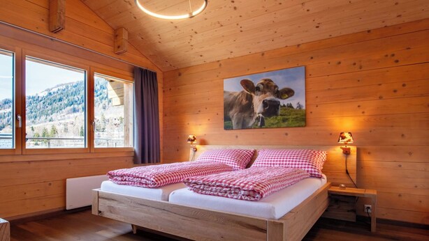 A wooden cabin bedroom with a bed, two bedside lamps, a window with curtains, and a cow painting on the wall.