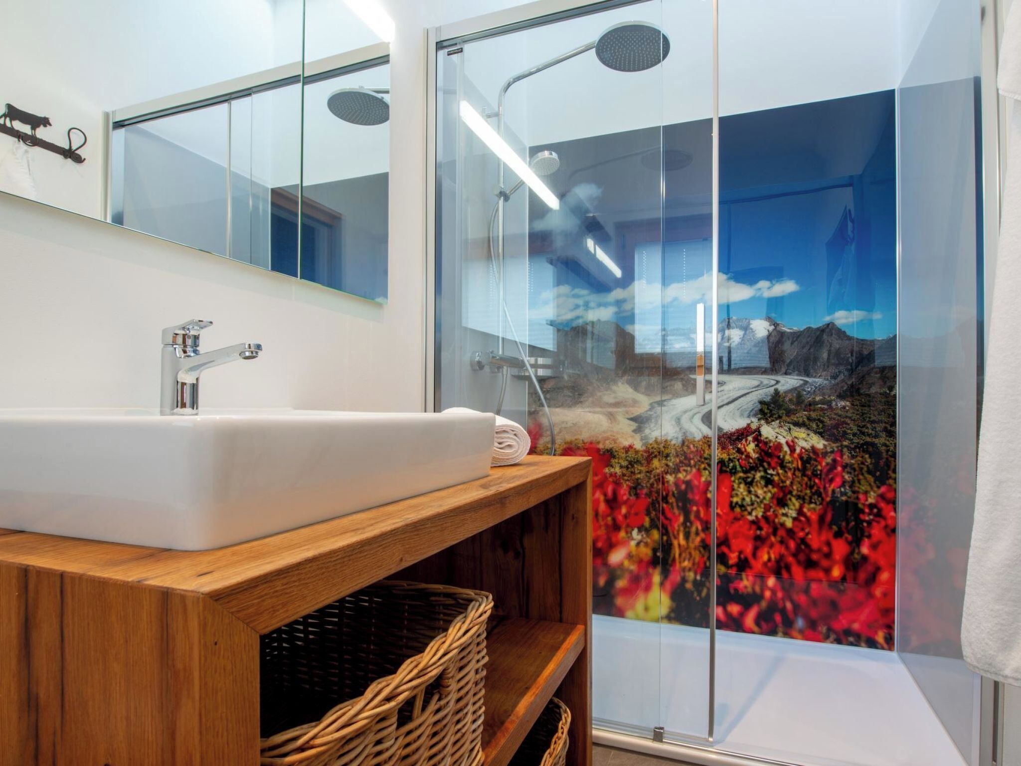 A modern bathroom with a large mirror, a white sink, a wooden vanity, and a glass shower enclosure with a scenic view.