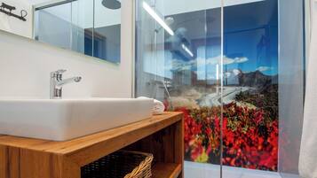 A modern bathroom with a large mirror, a white sink, a wooden vanity, and a glass shower enclosure with a scenic view.