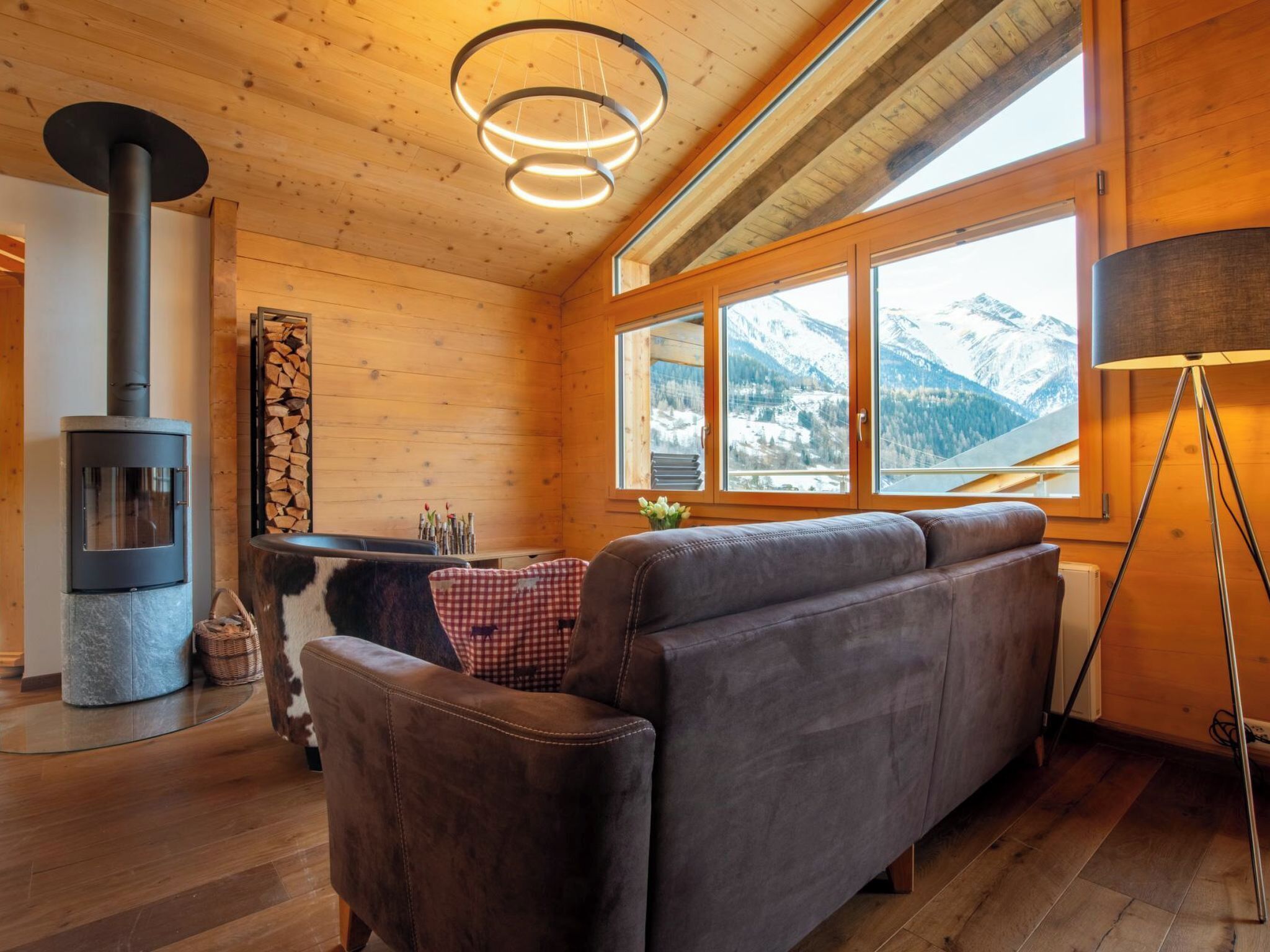 Ein gemütliches Blockhaus-Interieur mit Sofa, Kamin, Esstisch und Blick auf schneebedeckte Berge.