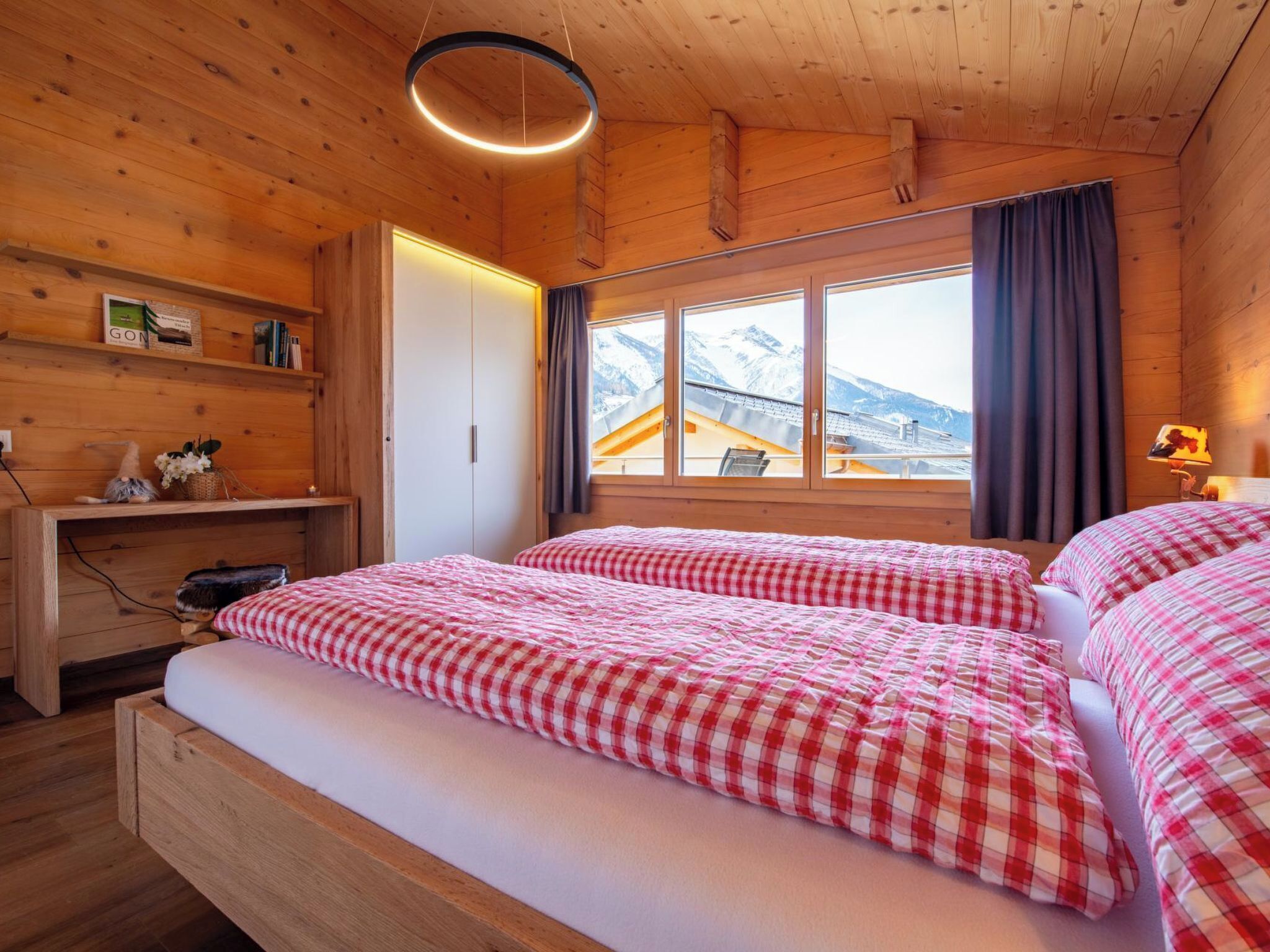 A wooden cabin bedroom with two beds, a window with a view of mountains, a shelf with books and a sign, and a hanging light fixture.