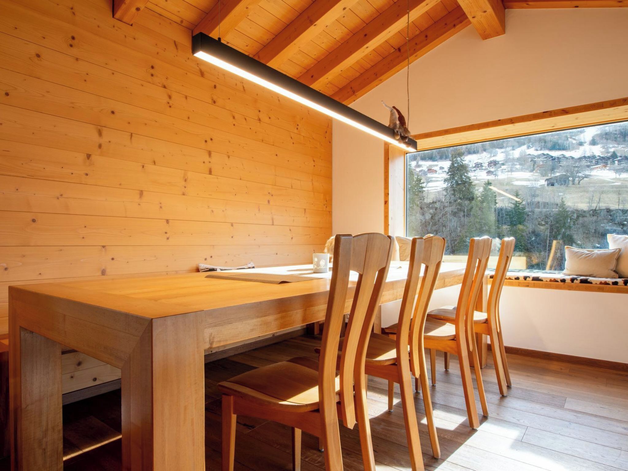 Ein Holzinterieur mit Esstisch, Stühlen, einem Fenster mit Aussicht und einer Pendelleuchte.
