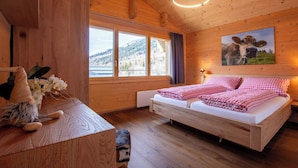 A wooden cabin bedroom with a bed, a nightstand, a gnome figure, a window with a view of a mountain, and a cow painting on the wall.