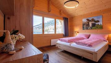 A wooden cabin bedroom with a bed, a nightstand, a gnome figure, a window with a view of a mountain, and a cow painting on the wall.