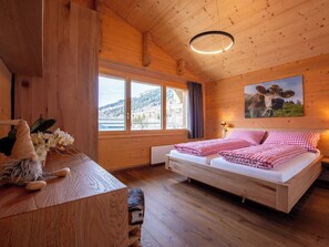 Un dormitori de cabana de fusta amb un llit, una tauleta de nit, una figura de gnom, una finestra amb vista a una muntanya i un quadre de vaca a la paret.