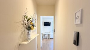 Un pasillo con una pared blanca, un estante pequeño con un florero, una puerta que da a una habitación con televisor y un termostato.