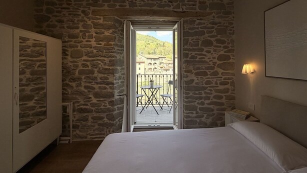 Un dormitorio con pared de piedra, cama, armario, balcón con sillas y vistas a edificios.