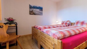 Una cama de madera individual con una colcha escocesa y una almohada roja, una silla de madera, una mesita con un jarrón de flores, y un cuadro de un camino sinuoso en la pared.