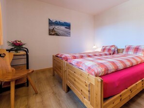 A single wooden bed with a plaid bedspread and a red pillow, a wooden chair, a small table with a vase of flowers, and a painting of a winding road on the wall. - Südlenz 13 1. OG Süd-West Appartement/Fewo, Bad, WC by Interhome (Riederalp)