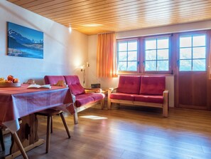Ein Zimmer mit Holzboden, einem Tisch mit rotem Tischläufer, einem roten Sofa und einem Fenster mit Vorhängen.
