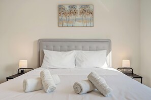 A neatly made bed with white linens and two rolled towels, a grey upholstered headboard, and a painting of trees on the wall. - Zeta's House, 550m from Agios Antonios Metro (Peristeri)