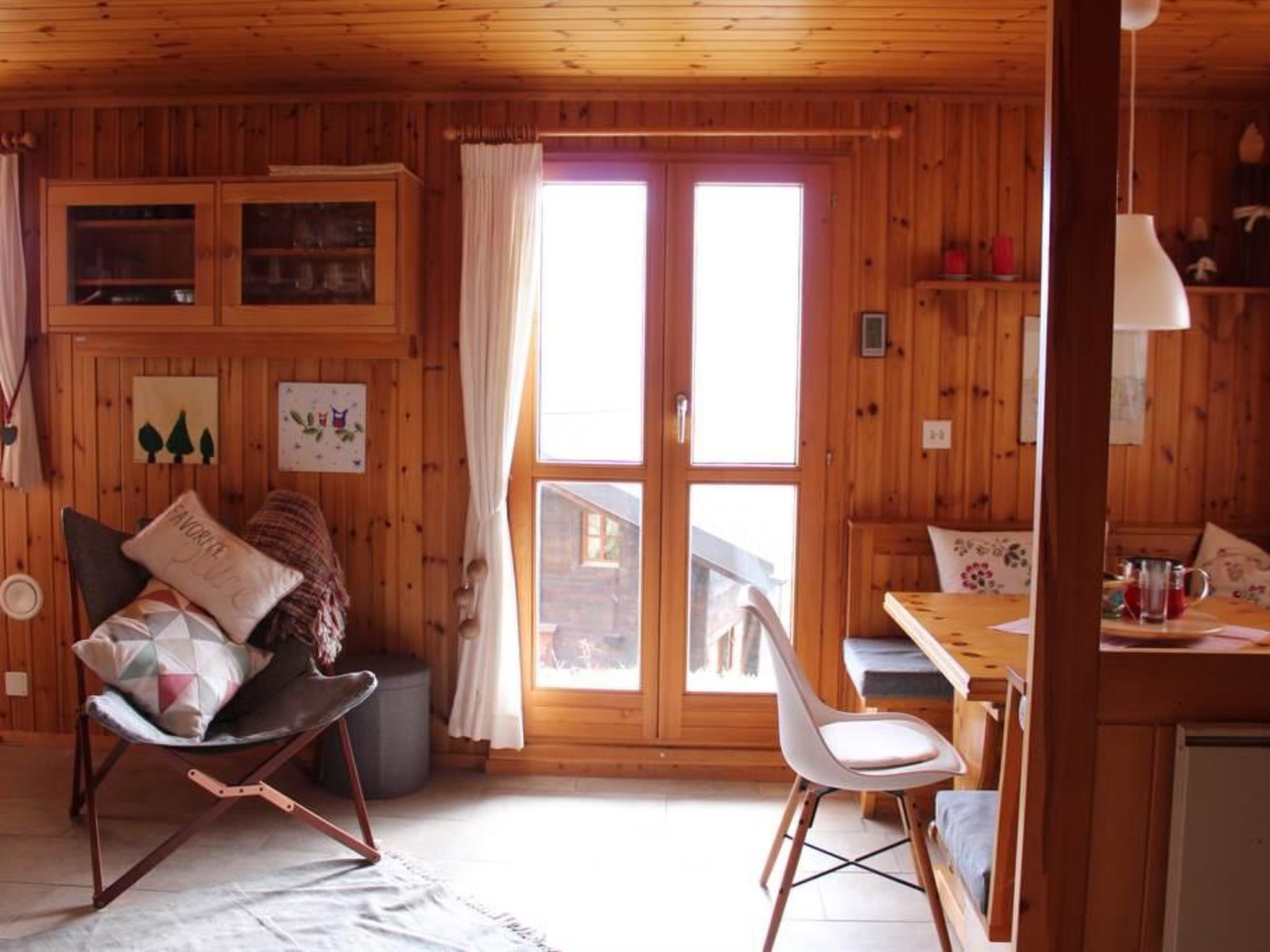 Interior d'una cabana de fusta amb zona de menjador, una cadira bressol i vista a través d'una porta de vidre.
