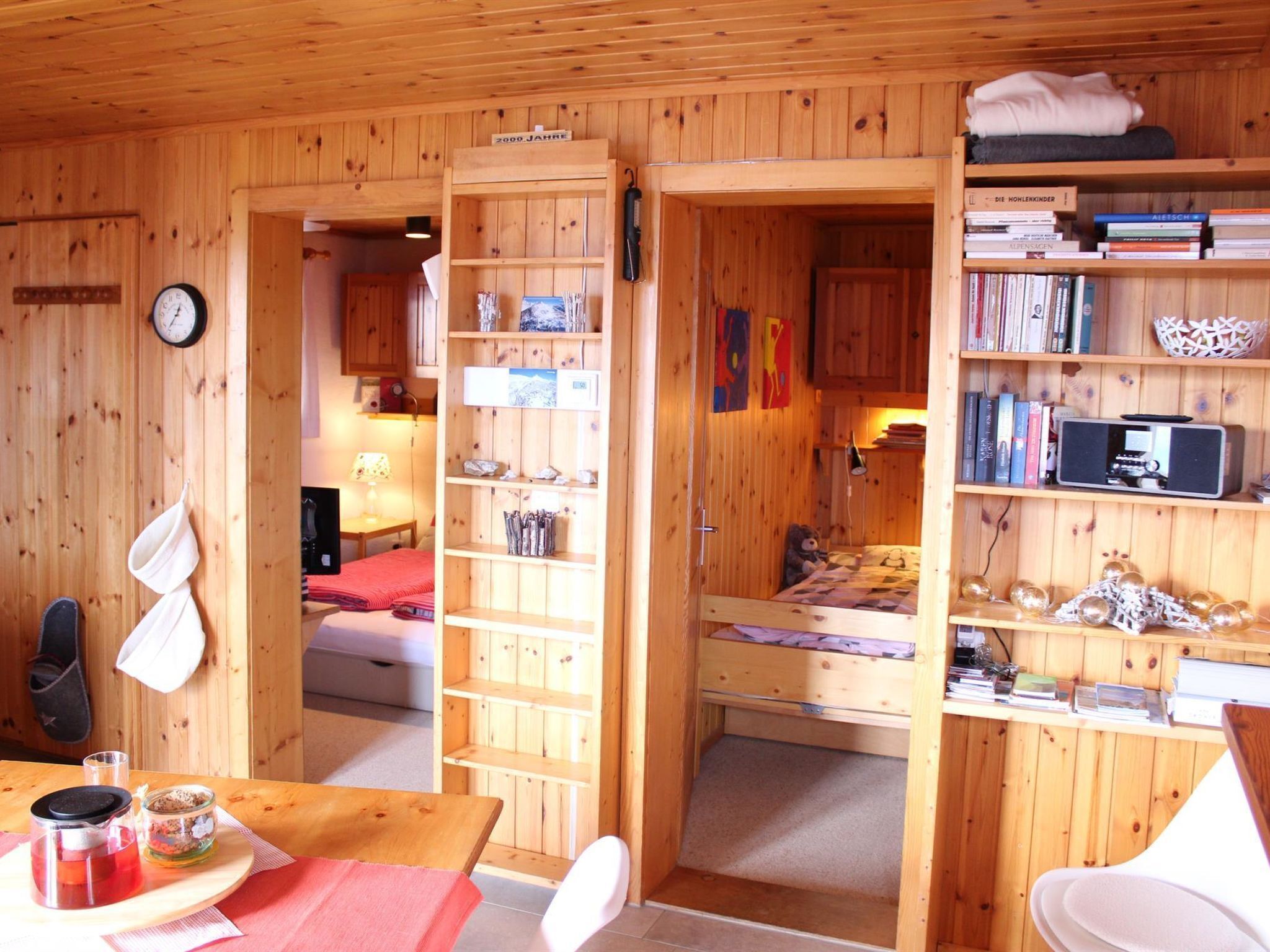 Ein Holzblockhaus-Interieur mit einem Bett, Regalen mit Büchern, einer Uhr und einem Tisch mit Teekanne.