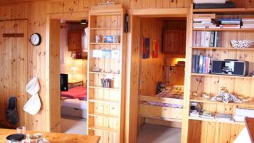 Ein Holzblockhaus-Interieur mit einem Bett, Regalen mit Büchern, einer Uhr und einem Tisch mit Teekanne.