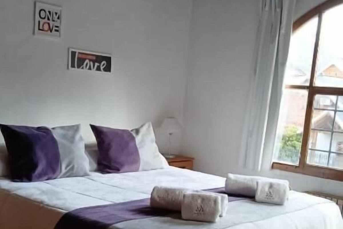 Een slaapkamer met een groot bed, houten balken, een raam met gordijnen en een muur met 'ON MY LOVE' decor.