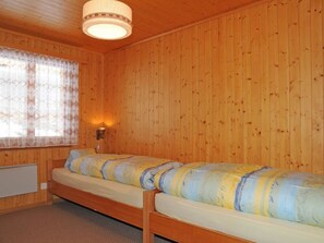 A room with wooden walls, a bed with patterned bedding, a window with lace curtains, and a wall-mounted light fixture.