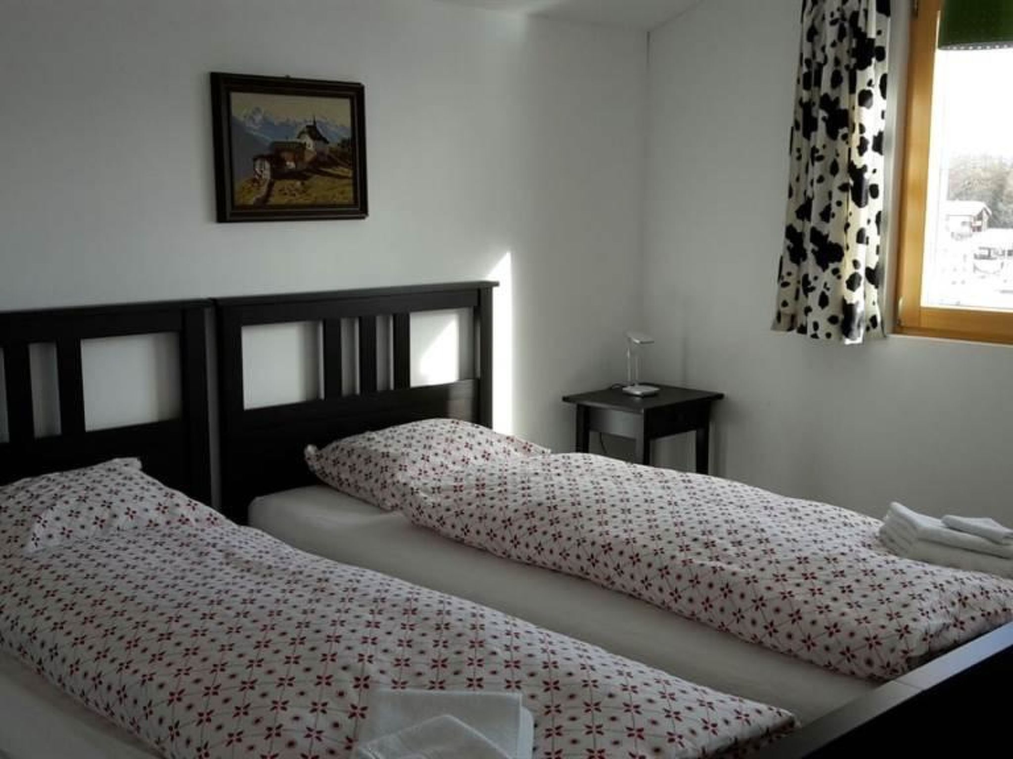 A room with two single beds, a small bedside table with a lamp, a framed picture on the wall, and a window with a curtain.