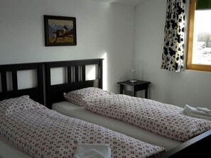 Una habitació amb dos llits individuals, una petita taula de nit amb una làmpada, un quadre amb marc a la paret i una finestra amb cortina.
