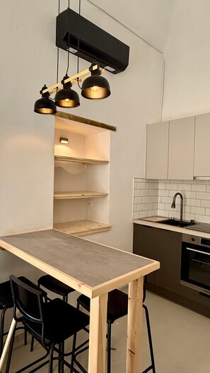 Una cocina con una mesa de madera, sillas negras, una lámpara colgante negra, salpicadero de azulejos blancos y un horno de acero inoxidable.